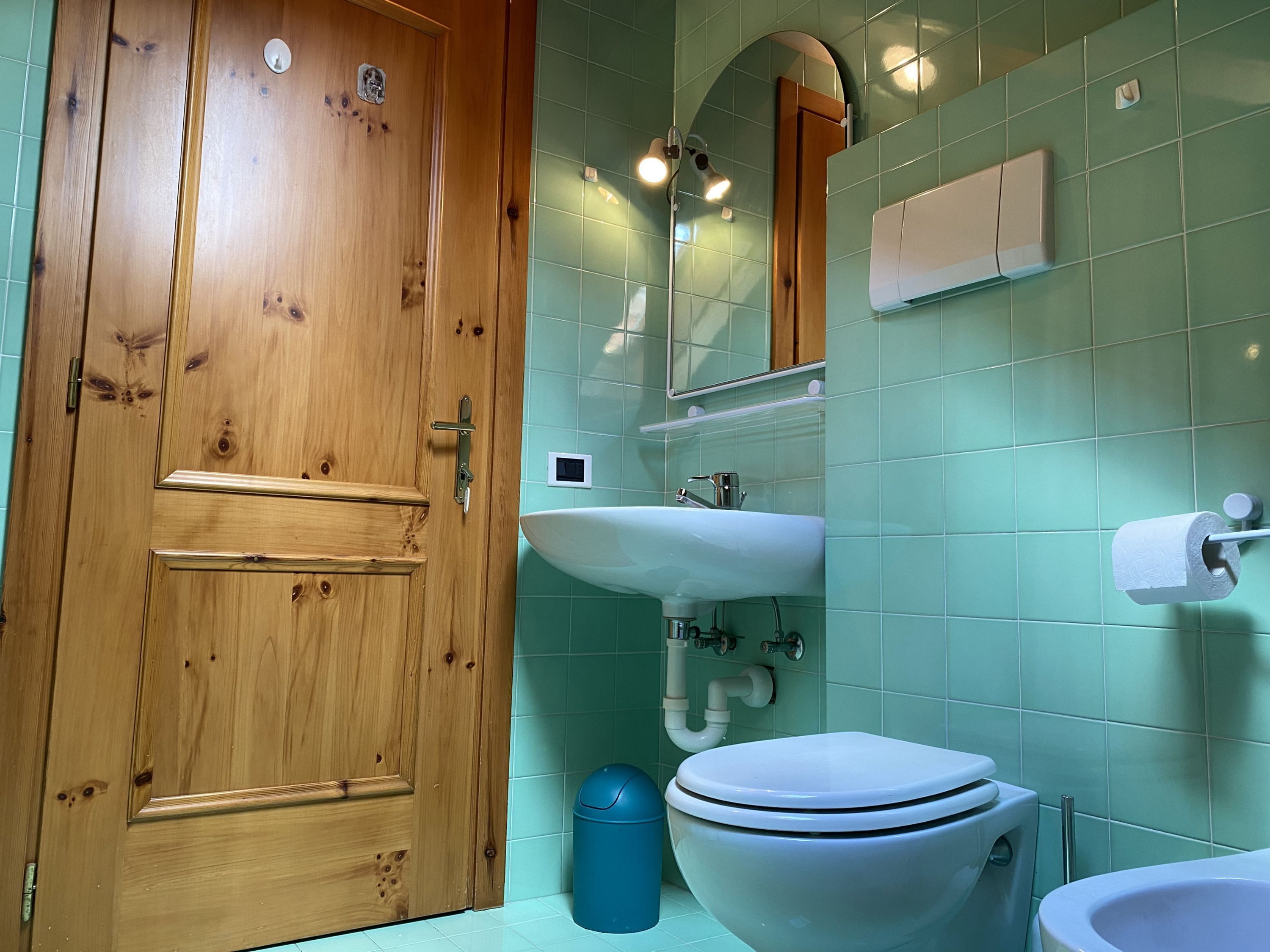 Un baño con puerta de madera, lavabo, inodoro y un cubo de basura azul.