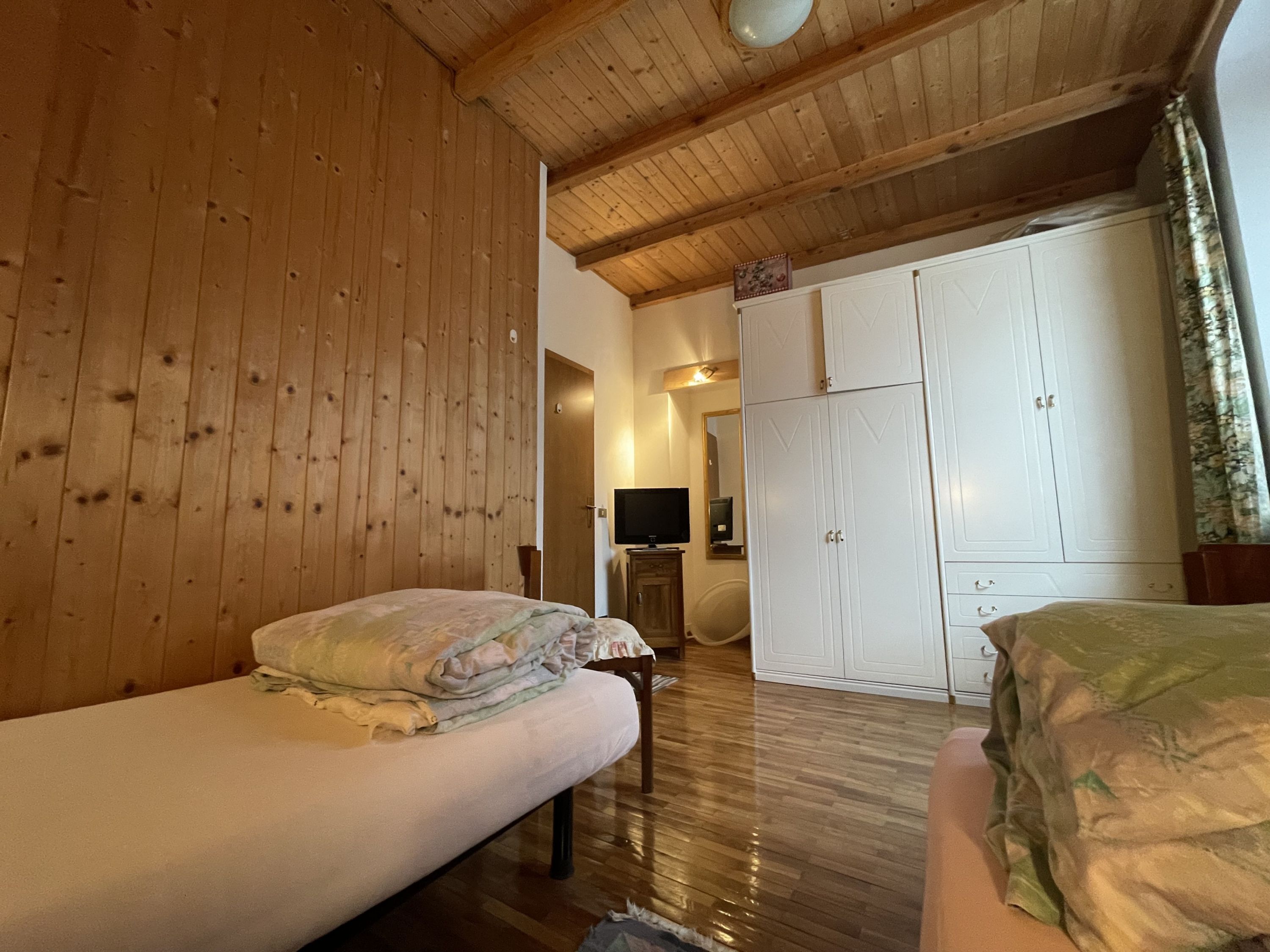 A room with wooden walls and ceiling, a bed with a blanket, a wardrobe, and a television.
