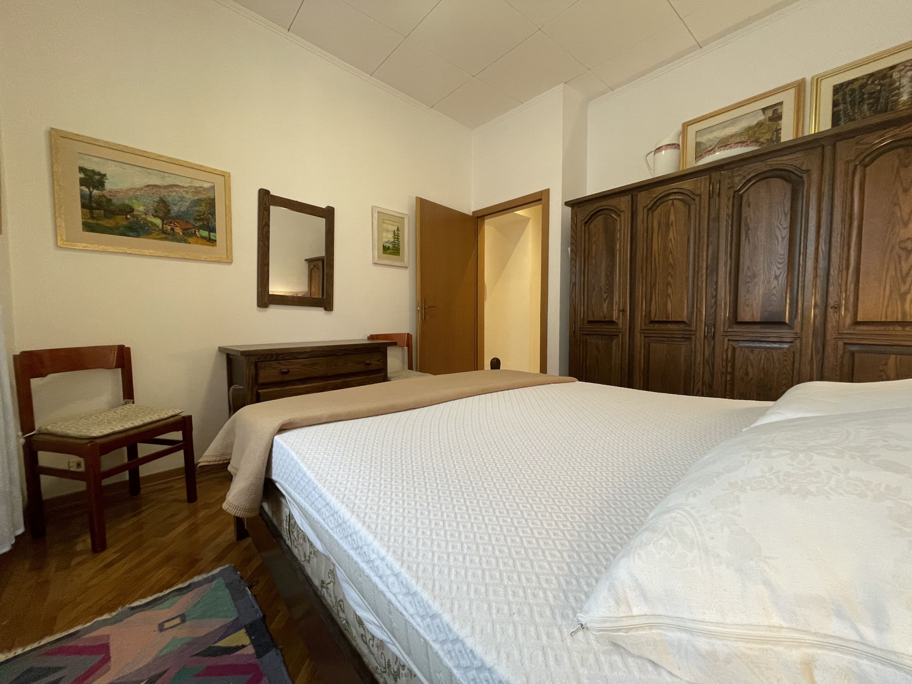 A bedroom with a bed, a wooden wardrobe, a chair, a dresser, and two framed pictures on the wall.