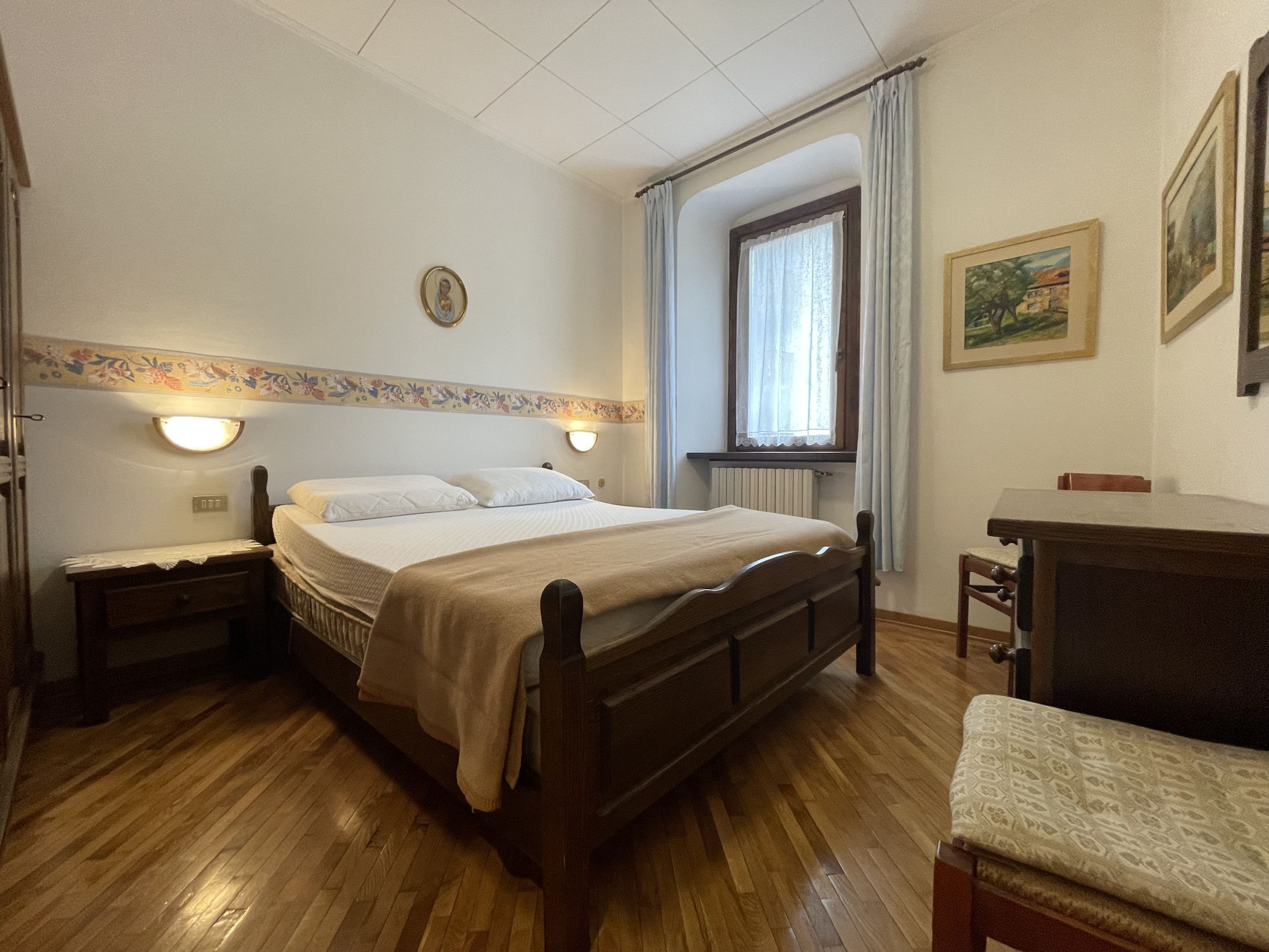 Ein Schlafzimmer mit einem hölzernen Bett, Nachttischen, einem kleinen Tisch, einem Fenster mit Vorhängen, einer Heizung und einem Schreibtisch mit Stuhl.