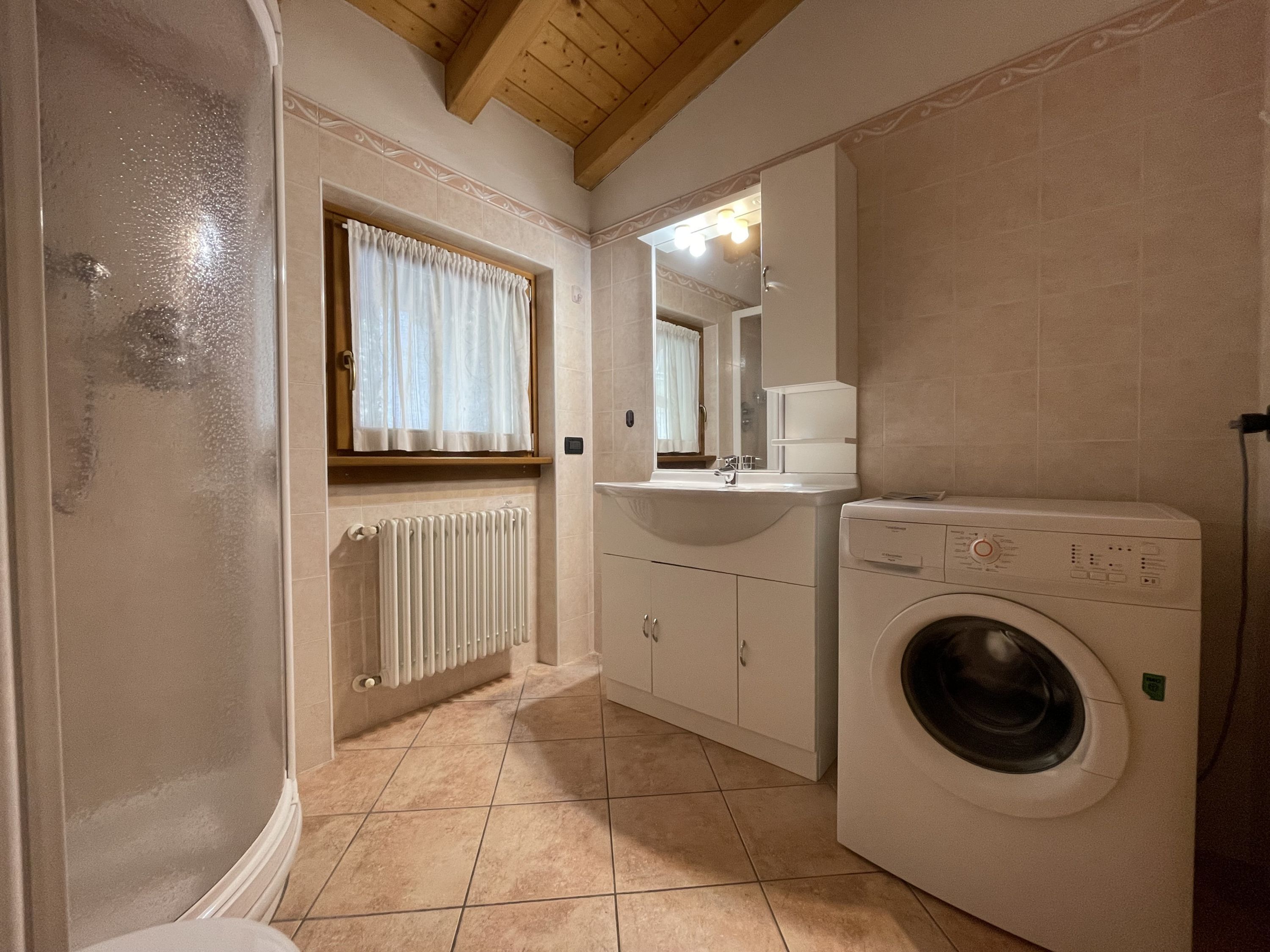 A compact bathroom with a built-in washing machine, a radiator, and a window with curtains.