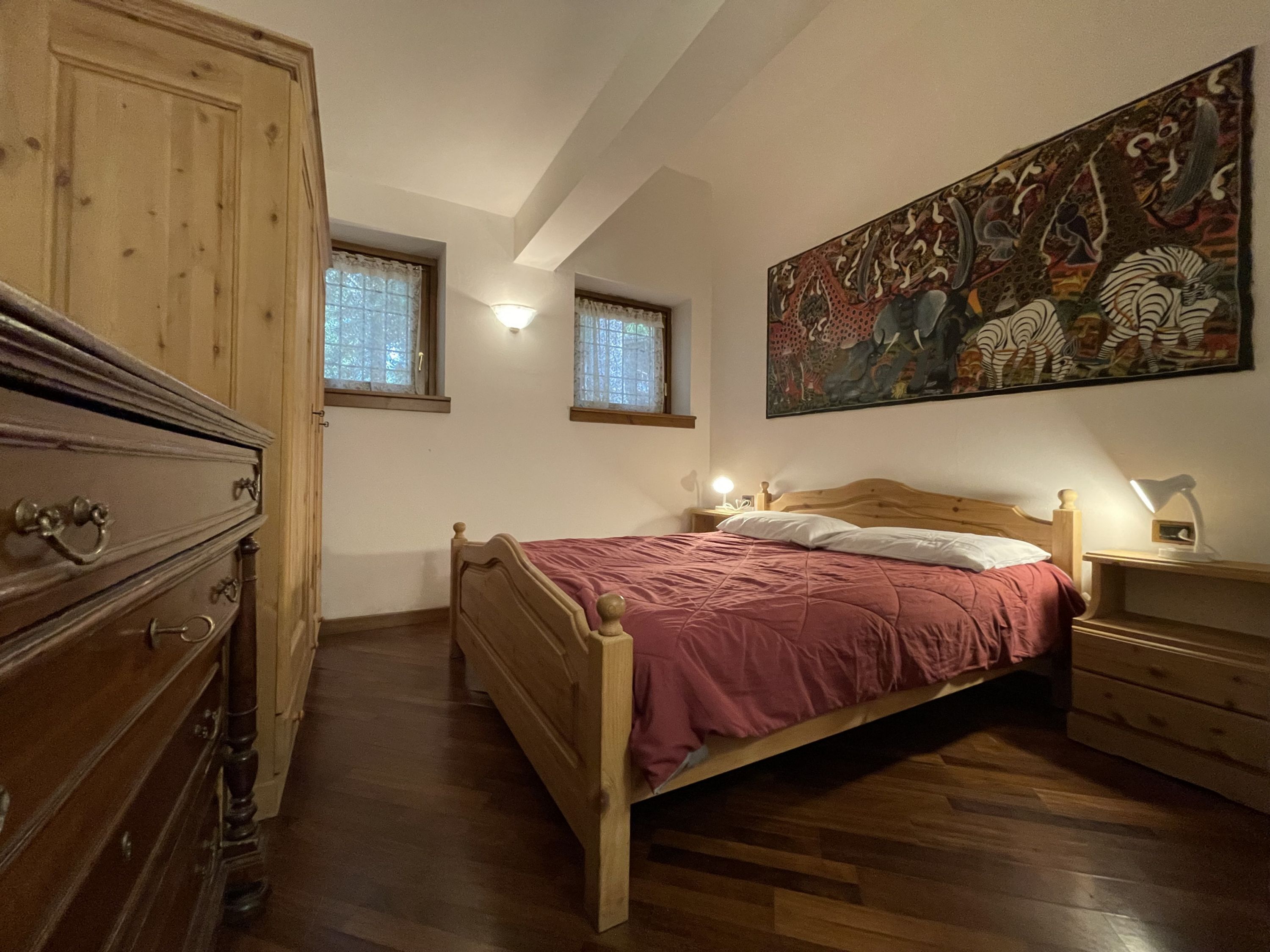 A bedroom with a wooden bed, a dresser, two windows with curtains, a wall painting, and a nightstand with a lamp.