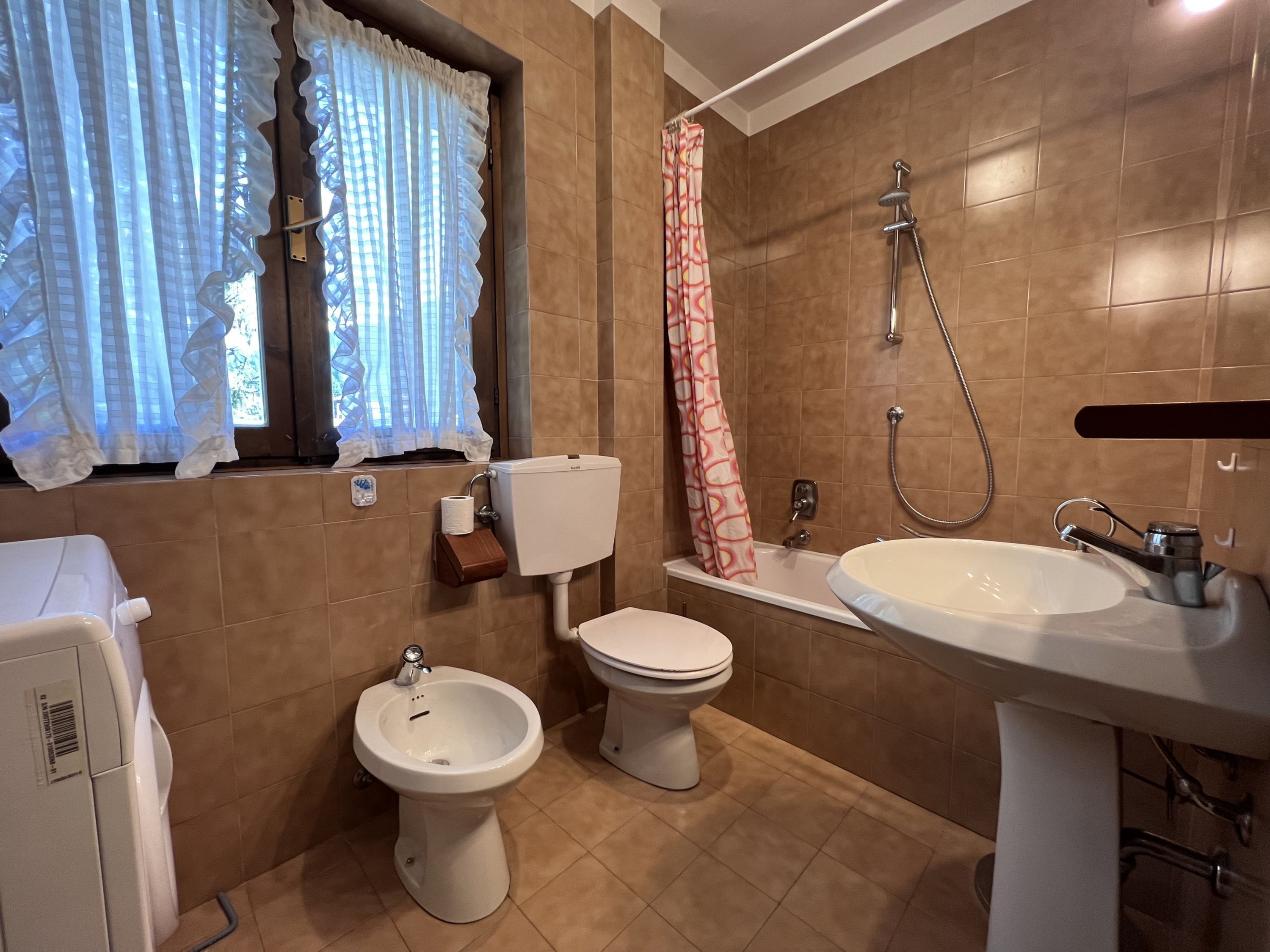Bagno con WC, bidet, lavabo, doccia e finestra con tende.