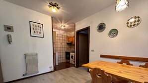 Zona pranzo con tavolo e sedie in legno, un termosifone, una lavatrice e una cucina sullo sfondo.