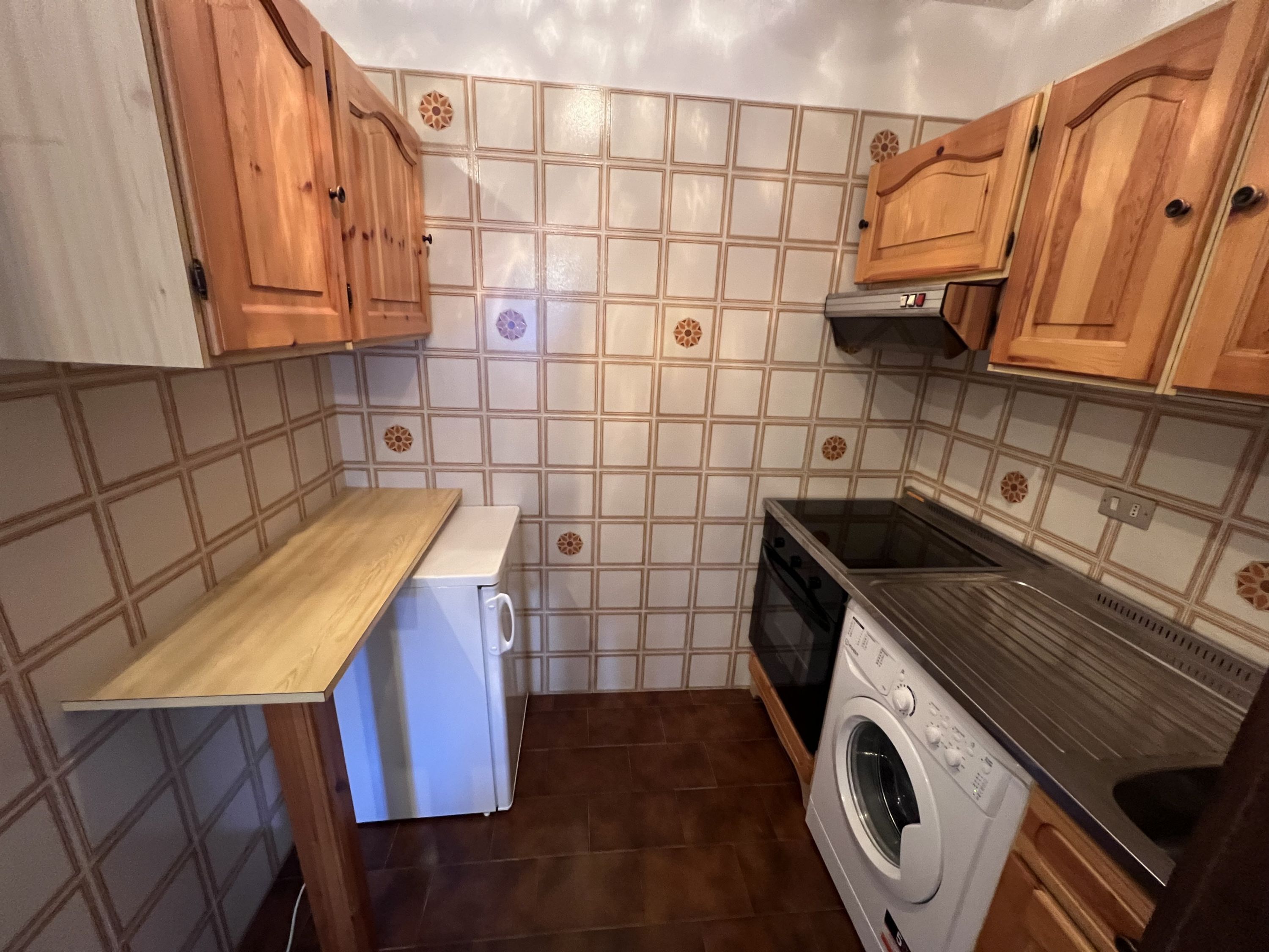 Een compacte keuken met houten kastjes, een inbouwoven, een wasmachine en een kleine koelkast.