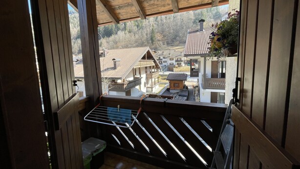 A wooden balcony with a metal drying rack, a small trash bin, and a view of a village with traditional houses. - 072 Trilocale Giustino (Giustino)