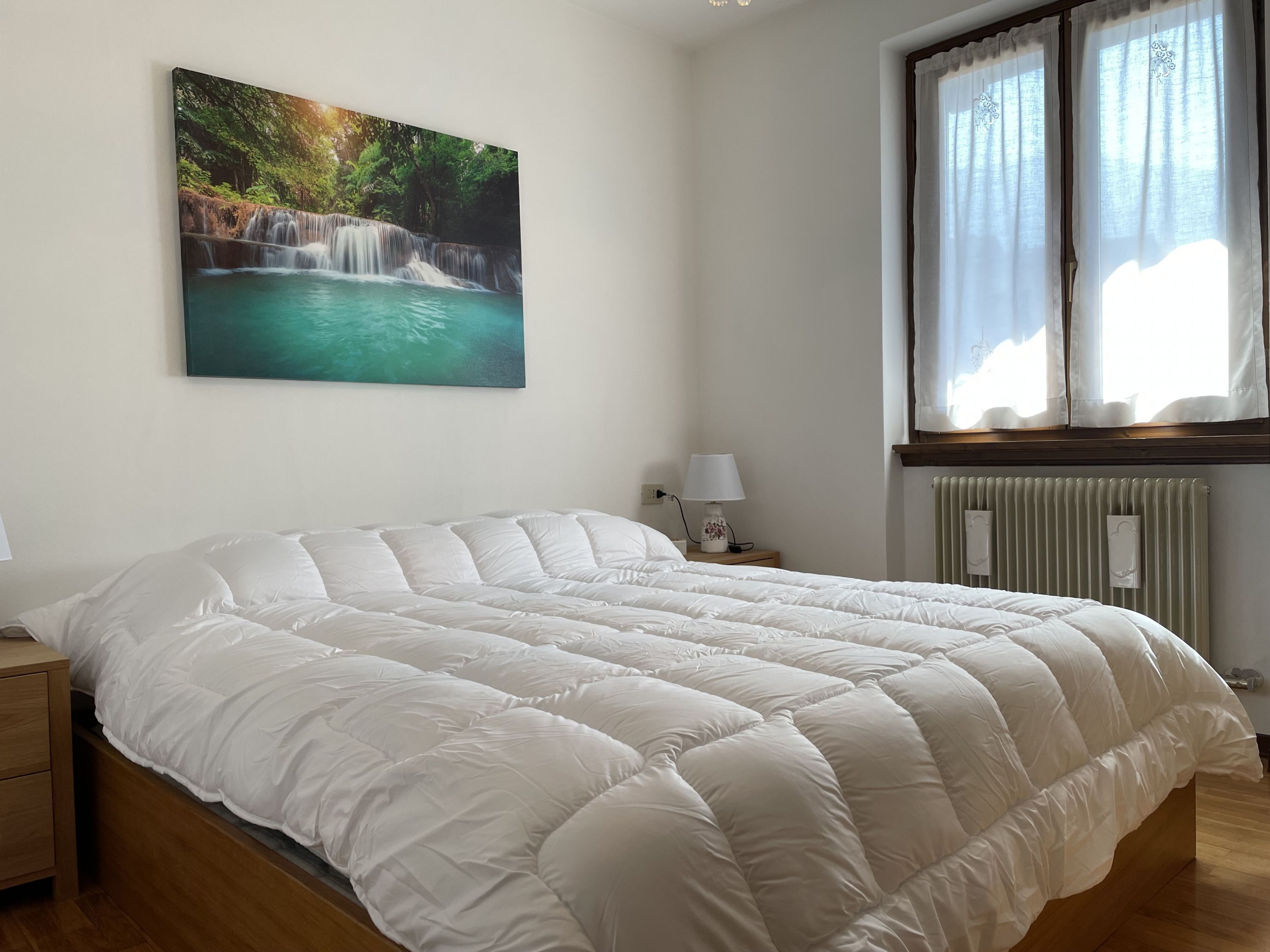 Una camera da letto con un letto grande, un comodino con una lampada, una finestra con tende e un quadro appeso al muro raffigurante una cascata.