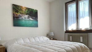 A bedroom with a large bed, a nightstand with a lamp, a window with curtains, and a wall-mounted painting of a waterfall. - 072 Trilocale Giustino (Giustino)