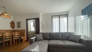 Una sala moderna con un sofá seccional gris, una mesa de comedor de madera con sillas, una pequeña cocina y un ventanal con cortinas.