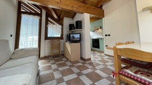 A cozy living room with a checkered floor, a wooden ceiling, a sofa, a television, and a kitchen area with light green cabinets. - 032 Luminosa Mansarda Pinzolo (Pinzolo)
