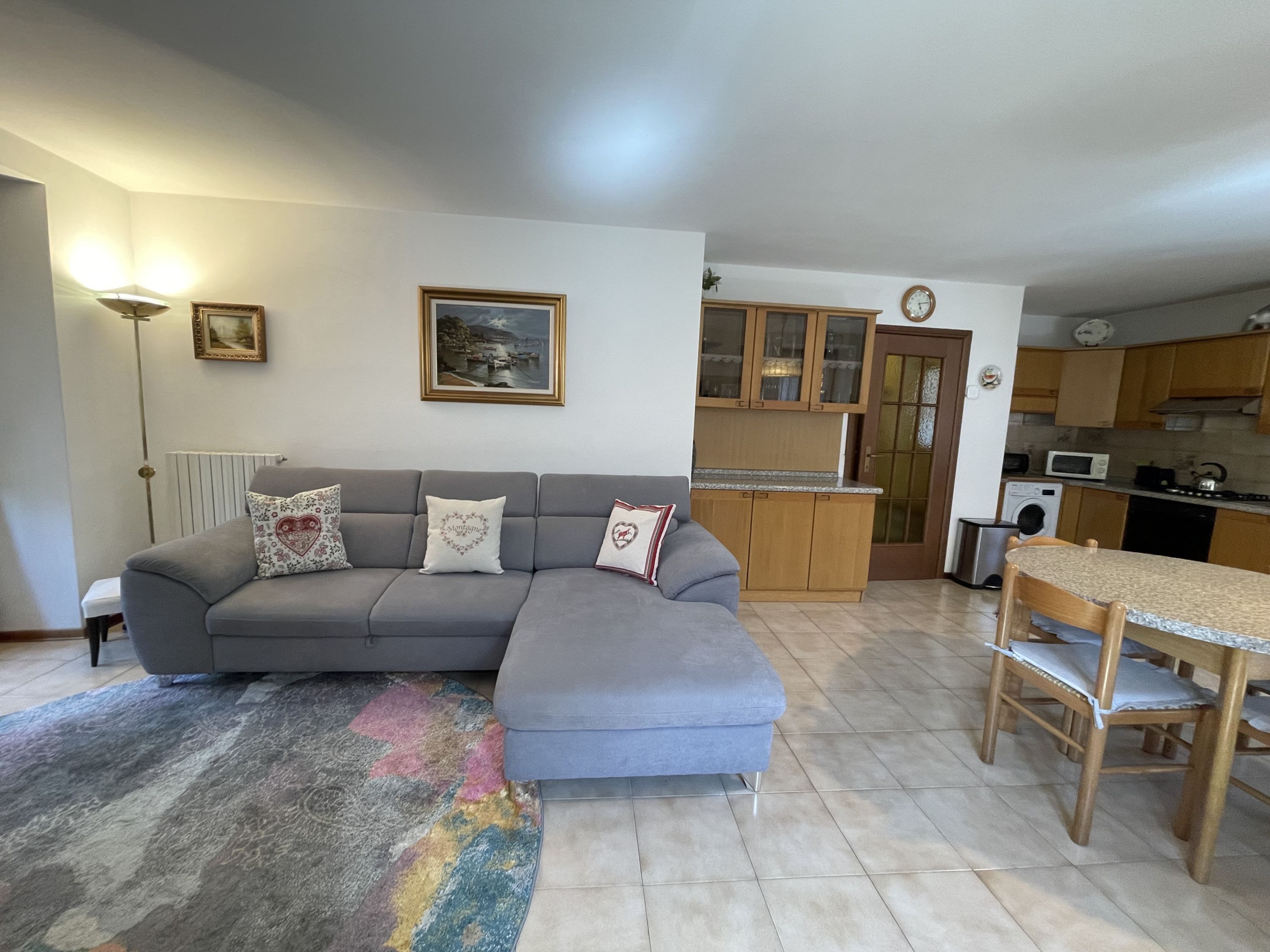 A living room with a grey sofa, a dining table with chairs, a kitchen area with wooden cabinets, and a radiator.