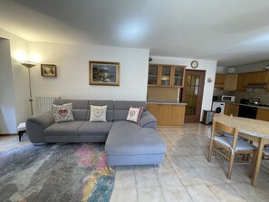 A living room with a grey sofa, a dining table with chairs, a kitchen area with wooden cabinets, and a radiator. - 062 Ampio Trilocale Carisolo (Carisolo)