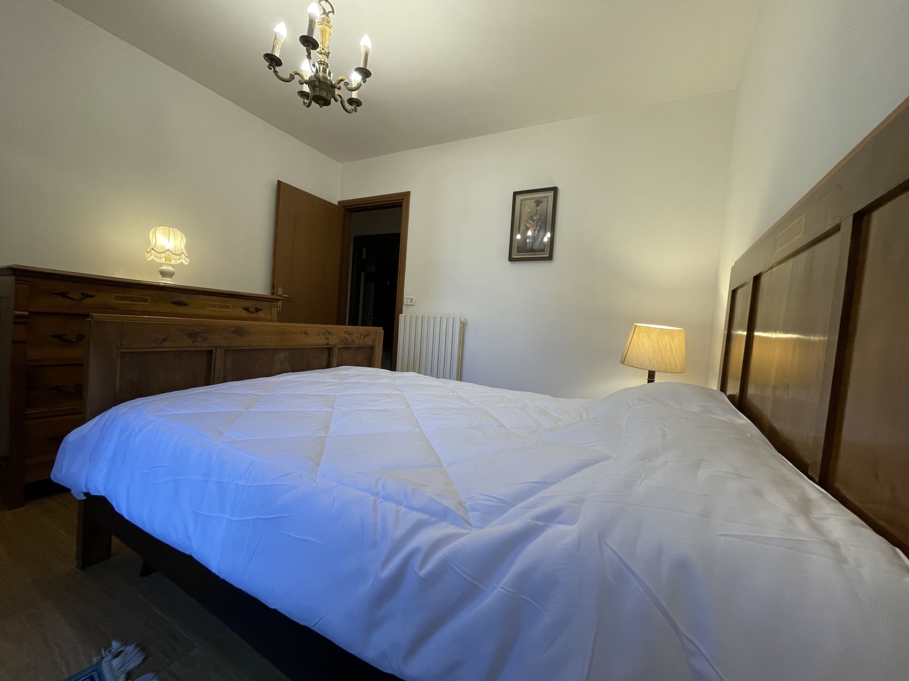 A bedroom with a large bed, wooden headboard, a dresser, a radiator, a framed picture on the wall, and a chandelier.