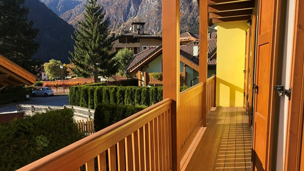 A wooden balcony with a tiled floor, leading to a yellow door. In the background, there are houses with sloped roofs and a mountain range. - 012 Trilocale a due Passi Dagli Impianti Pinzolo (Carisolo)