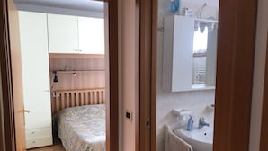 A bedroom with a bed, wooden headboard, and a wardrobe. Adjacent is a bathroom with a sink, mirror, and towel rack. - 012 Trilocale a due Passi Dagli Impianti Pinzolo (Carisolo)