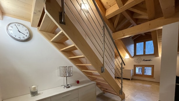 A spacious room with a wooden staircase, a white wall clock, a desk with drawers, a lamp, and a window with a view. - 061 Bilocale Mansarda Carisolo (Carisolo)