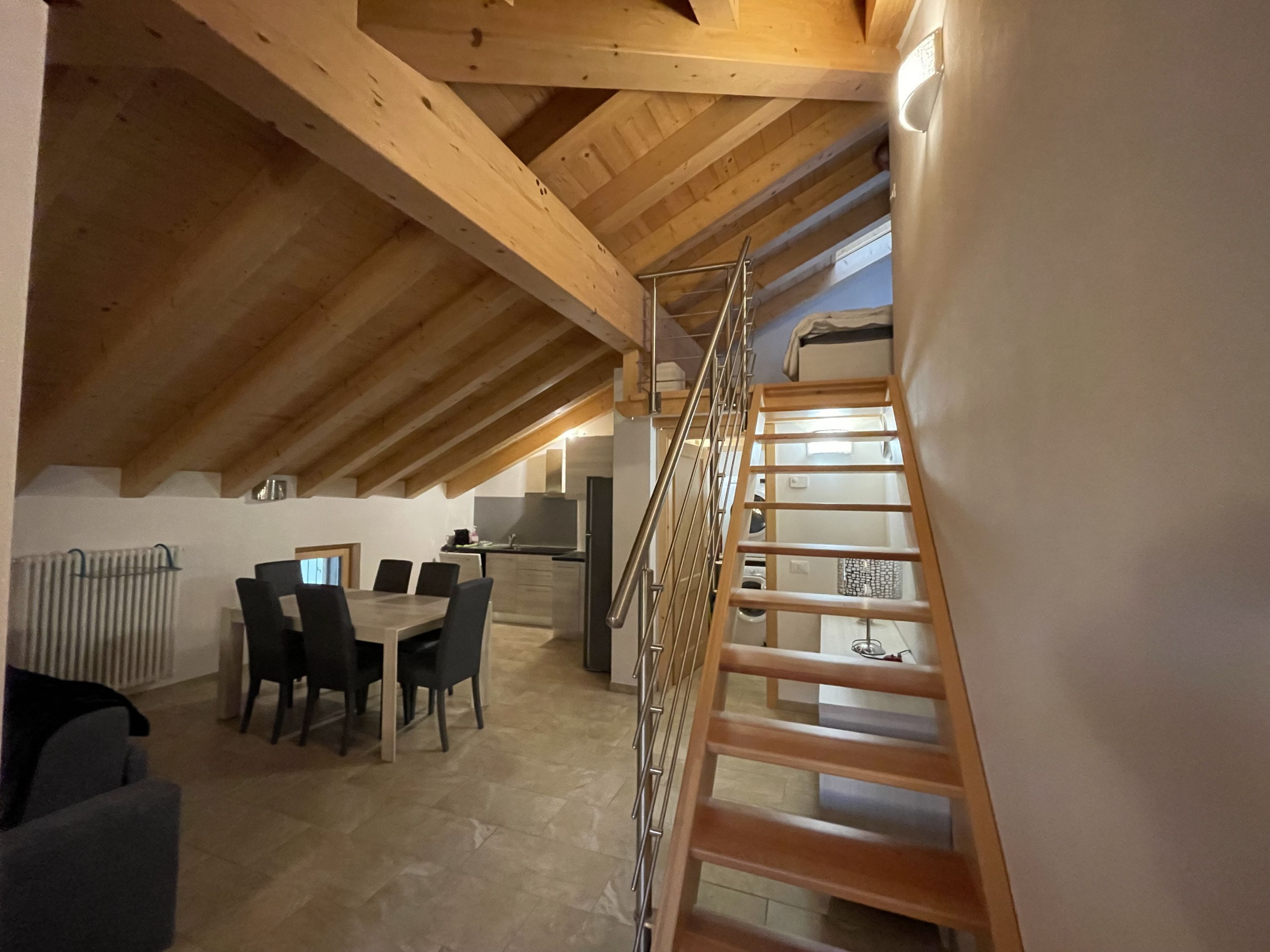 An attic bedroom with a wooden ceiling, a staircase, a dining area with a table and chairs, and a kitchen in the background.