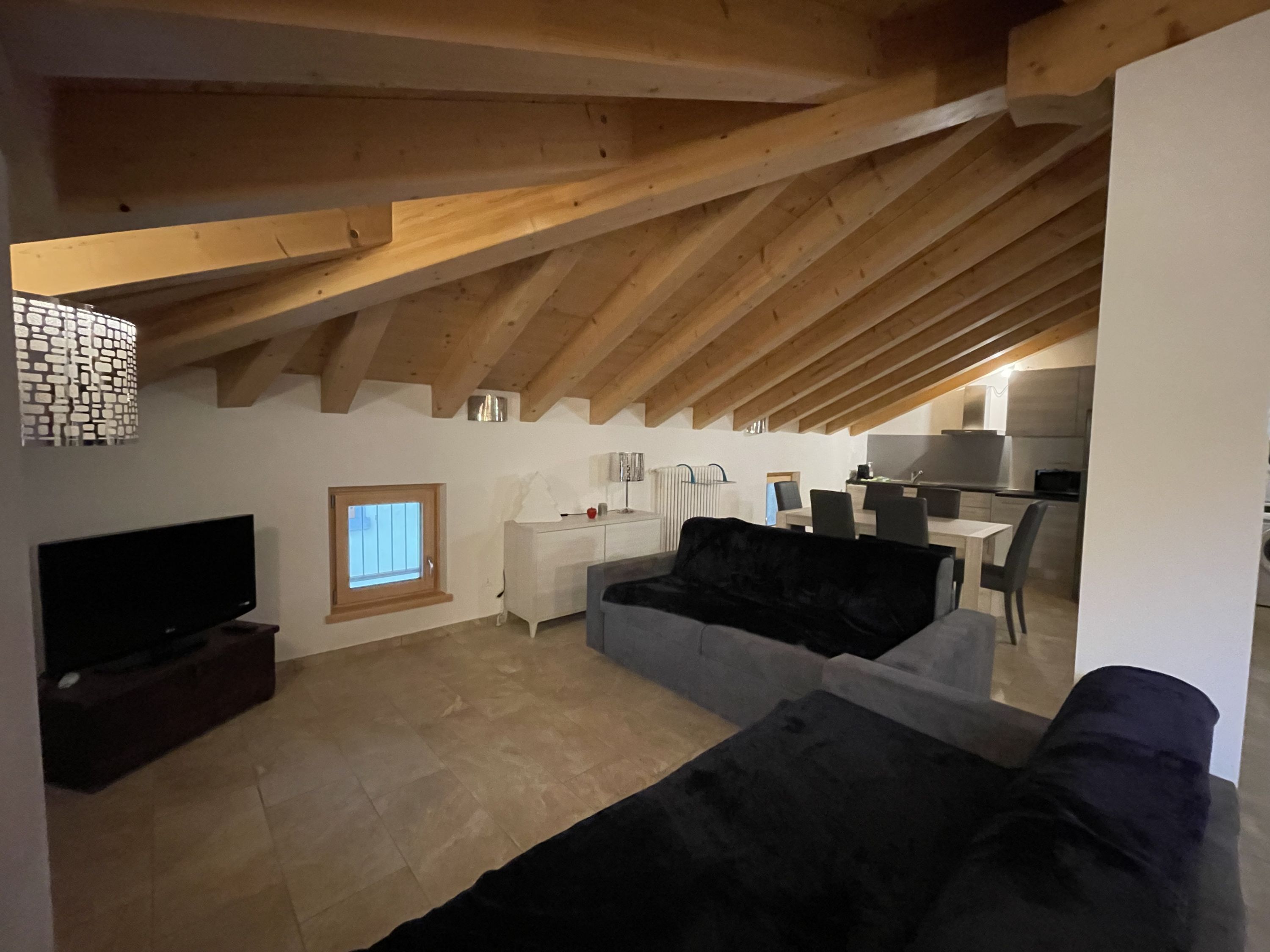 A loft-style living space with exposed wooden beams, a flat-screen TV, a sofa, and a dining area with chairs and a table.