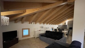 A loft-style living space with exposed wooden beams, a flat-screen TV, a sofa, and a dining area with chairs and a table. - 061 Bilocale Mansarda Carisolo (Carisolo)