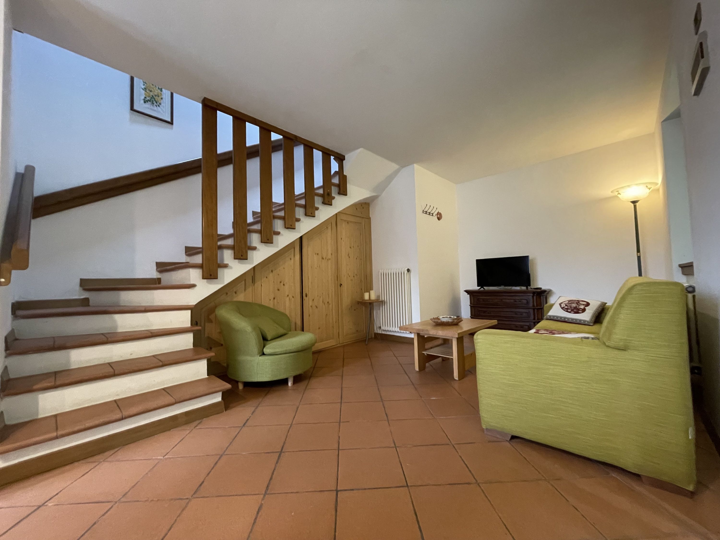 Un salon avec un escalier, un fauteuil vert, un meuble en bois et un téléviseur.