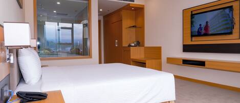 Habitación de hotel con una cama grande, un escritorio con teléfono, un televisor montado en la pared y vistas a la ciudad a través de un ventanal amplio.