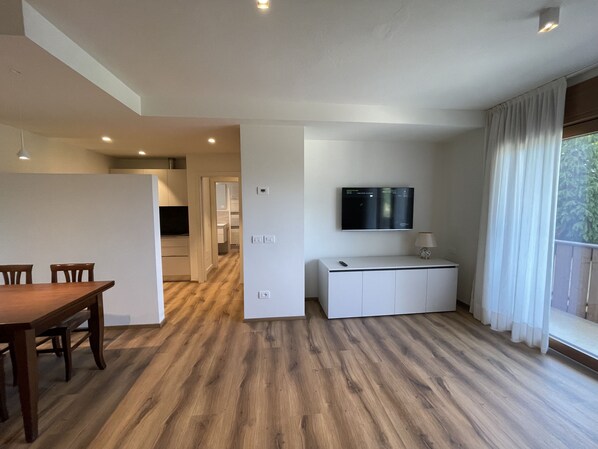 Ein modernes Wohnzimmer mit Holzboden, einem Esstisch, einem an der Wand befestigten Fernseher und einem Balkon mit Vorhängen.