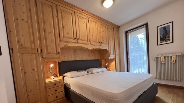 Ein Schlafzimmer mit Holzschrank, Bett mit weißer Bettwäsche, Nachttisch mit Lampe und Heizkörper.