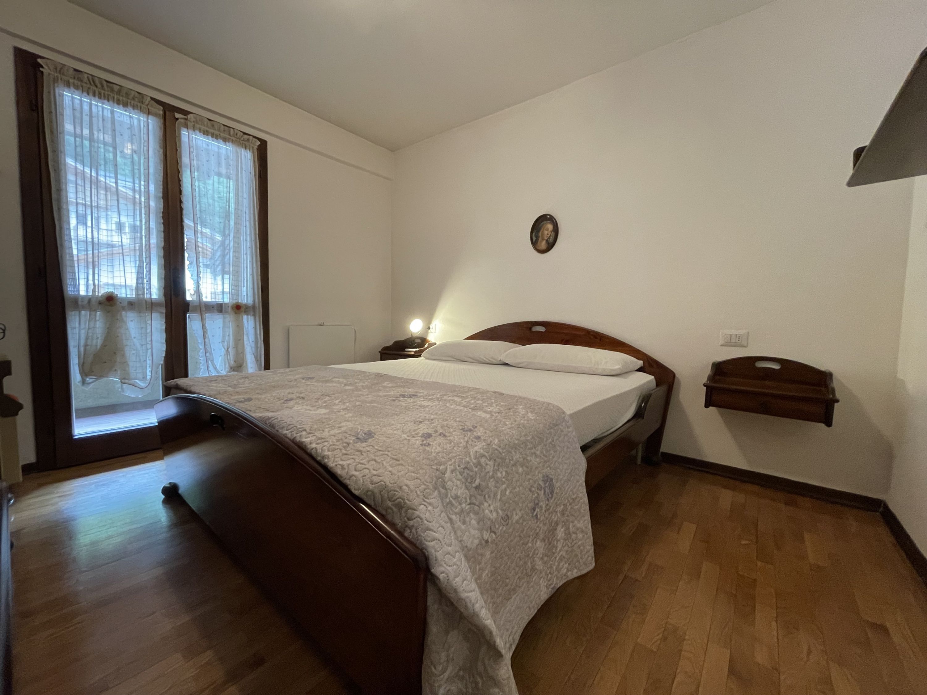 A bedroom with a wooden bed, a bedside table with a lamp, a small wooden shelf, and a window with curtains.