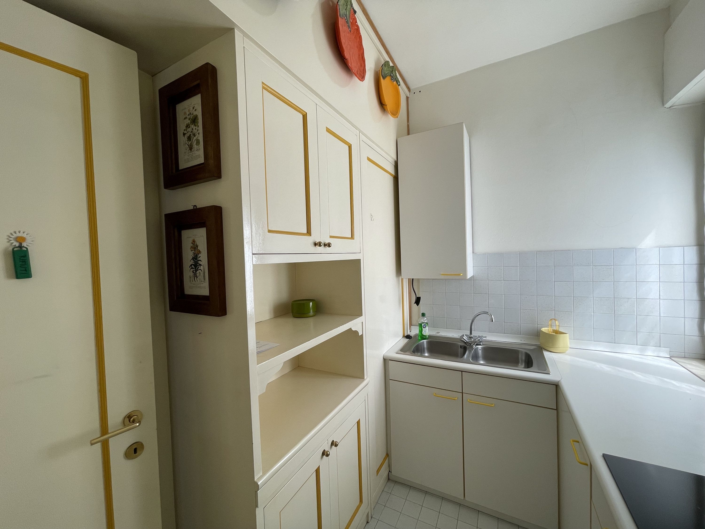 Une cuisine avec des meubles blancs, un évier et un plan de travail. Des tableaux encadrés ornent le mur et un bol vert est posé sur une étagère.