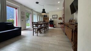 A spacious living area with a dining table and chairs, a black sofa, and a kitchen area with wooden cabinets and a TV. - 067 Bilocale Piano Terra Giustino (Giustino)