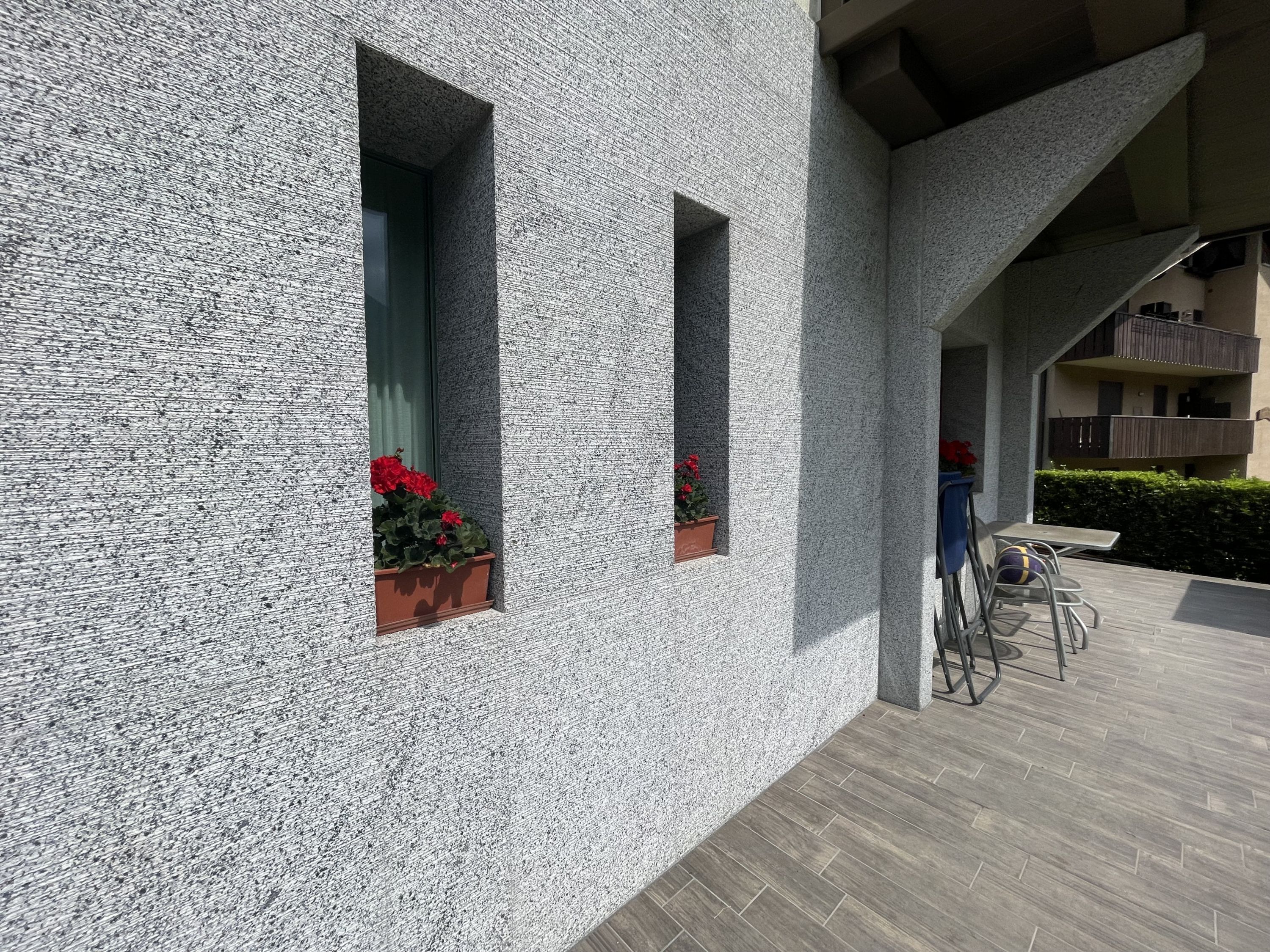 Un edificio moderno con paredes exteriores texturizadas, plantas en macetas en jardineras y un espacio al aire libre cubierto con una mesa y sillas.