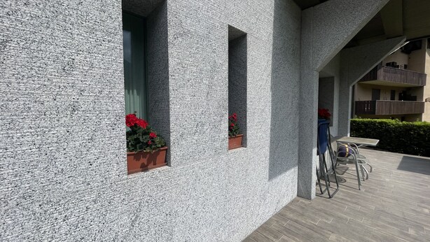 Ein modernes Gebäude mit strukturierten Außenwänden, Blumentöpfen in Fensterkästen und einer überdachten Außenbereich mit Tisch und Stühlen.