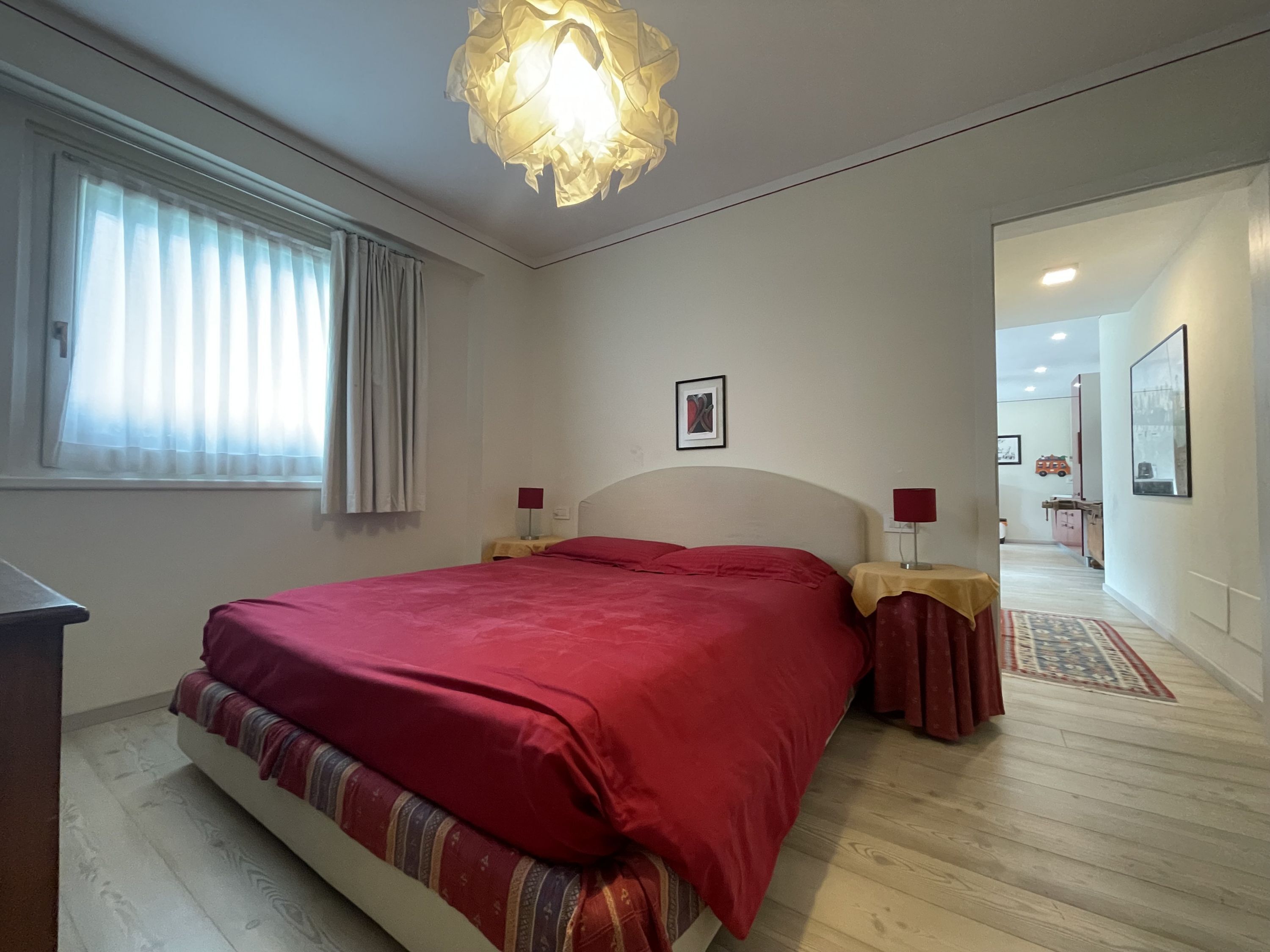 Ein Schlafzimmer mit einem Bett, zwei Nachttischen, einer Fensterbank, einem Vorhangfenster, einer Hängeleuchte und einer Tür zu einem anderen Raum.