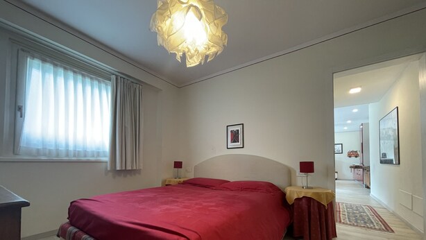 Une chambre à coucher comprenant un lit, deux tables de chevet, une commode, une fenêtre avec des rideaux, un luminaire suspendu et une porte donnant sur une autre pièce.