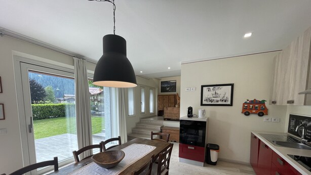 A dining area with a wooden table and chairs, a black pendant light, and a view of a garden through large windows. - 067 Bilocale Piano Terra Giustino (Giustino)