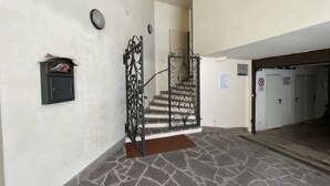 A hallway with a stone floor, a mailbox, a staircase with a wrought iron railing, and a door with a '10' sign. - 079 Bilocale Pinzolo (Pinzolo)