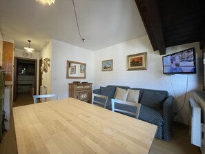 A cozy living room with a wooden dining table, a patterned sofa, and a television mounted on the wall. - 079 Bilocale Pinzolo (Pinzolo)