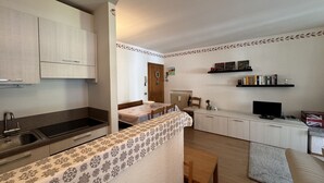 Eine kompakte Küche mit Spüle, Schränken und einem Essbereich mit Tisch und Stühlen. Ein kleiner Fernseher und ein Regal mit Büchern und Dekorationen sind ebenfalls vorhanden.