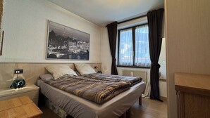 Ein Schlafzimmer mit einem Bett, einem Nachttisch, einem Holztisch, einem Fenster mit Vorhängen und einem Stadtbild an der Wand.