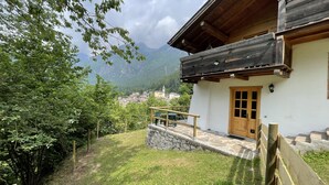 A chalet with a stone foundation, wooden balcony, and a view of a village and mountains in the background.