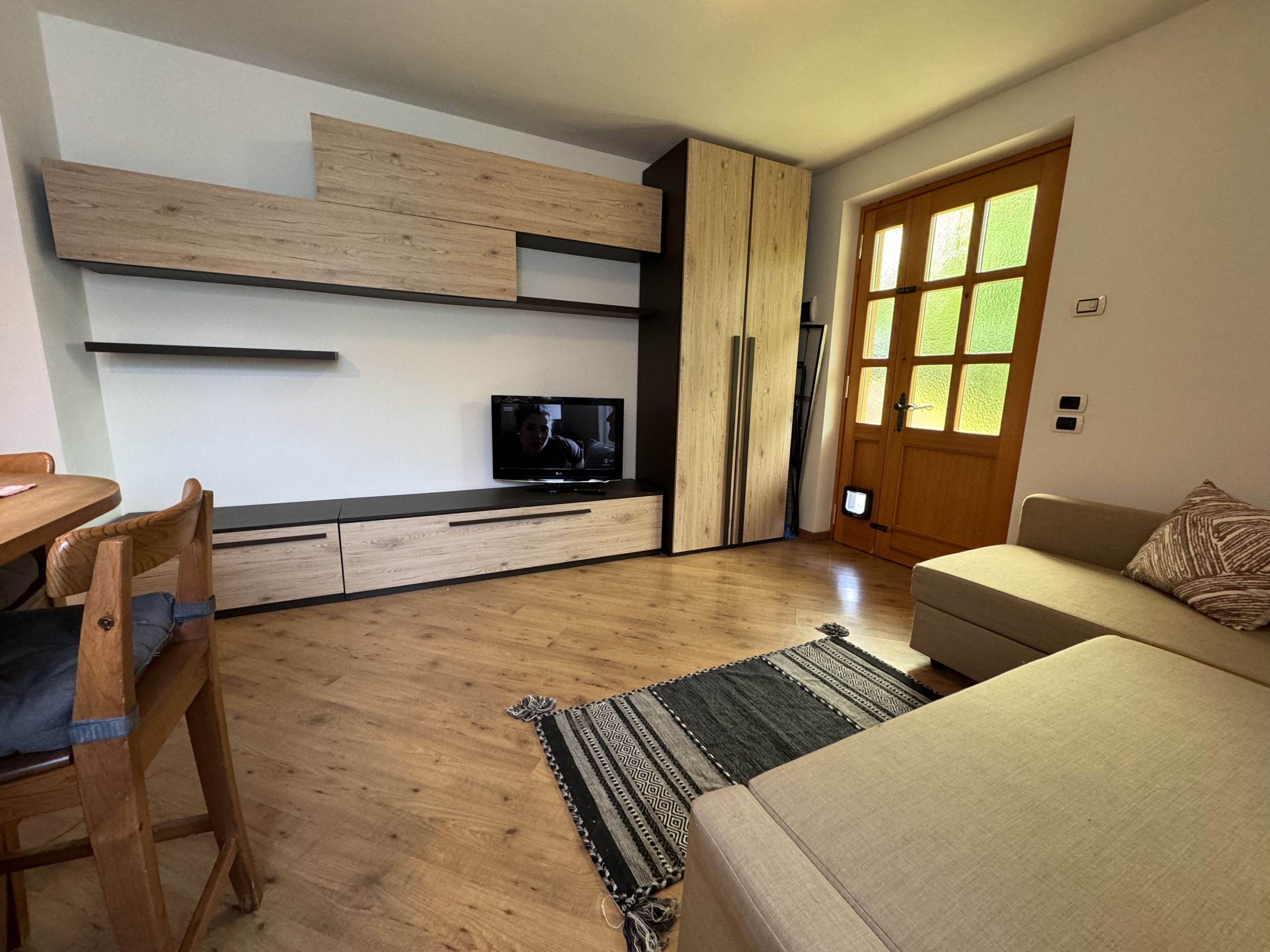 Ein modernes Wohnzimmer mit Holzboden, einer Couch, einem Fernseher auf einem Holzschrank, einem Esstisch mit Stühlen und einem großen Fenster mit Blick auf Grünflächen.