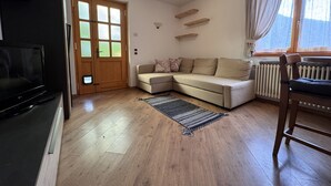 A living room with a beige sectional sofa, a television, a wooden floor, a window with curtains, and a radiator.