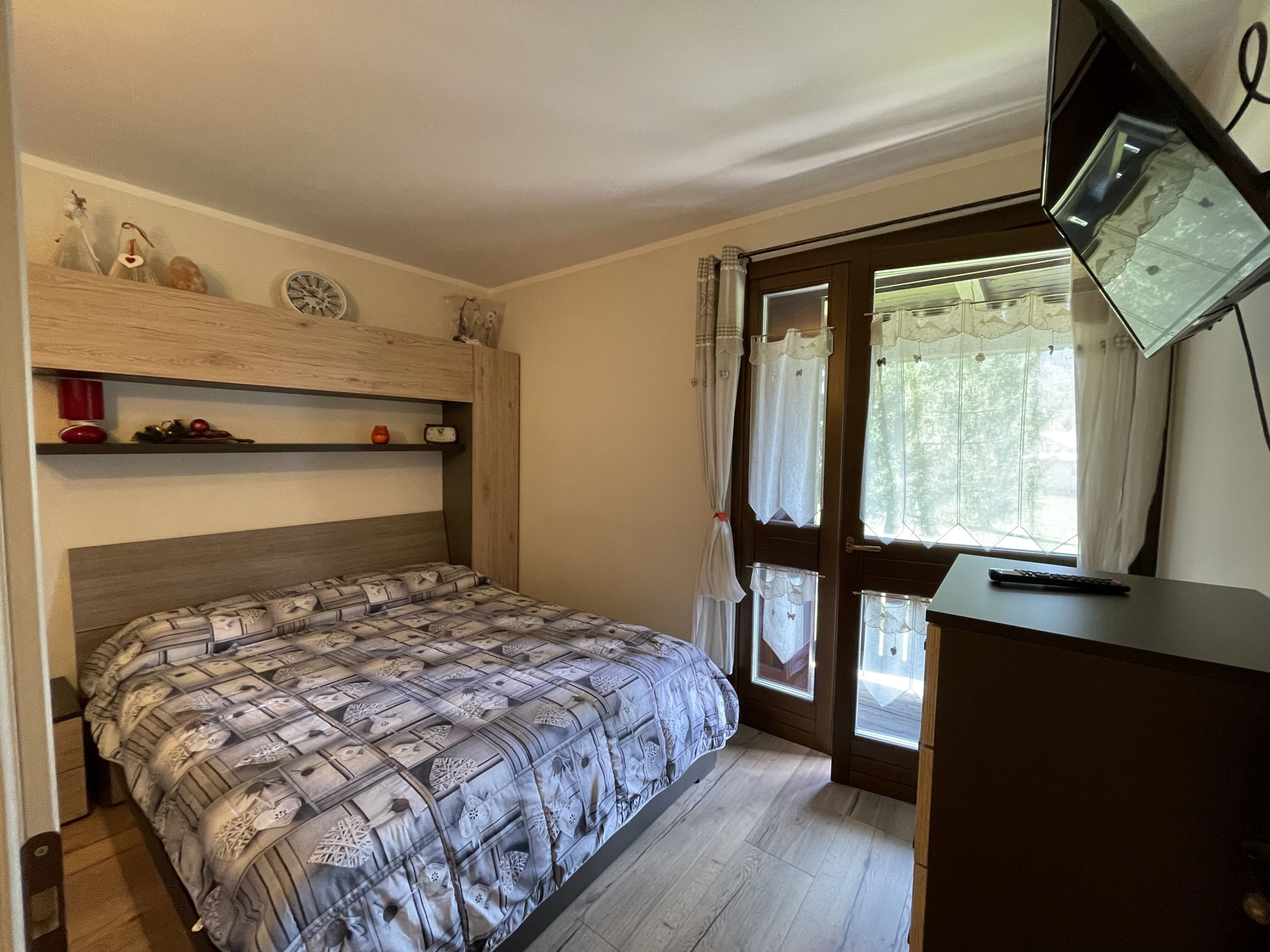 A bedroom with a bed, a wooden headboard, a shelf with decorative items, a window with curtains, and a desk with a lamp.