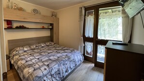 A bedroom with a bed, a wooden headboard, a shelf with decorative items, a window with curtains, and a desk with a lamp. - 082 Bilocale Pinzolo (Pinzolo)