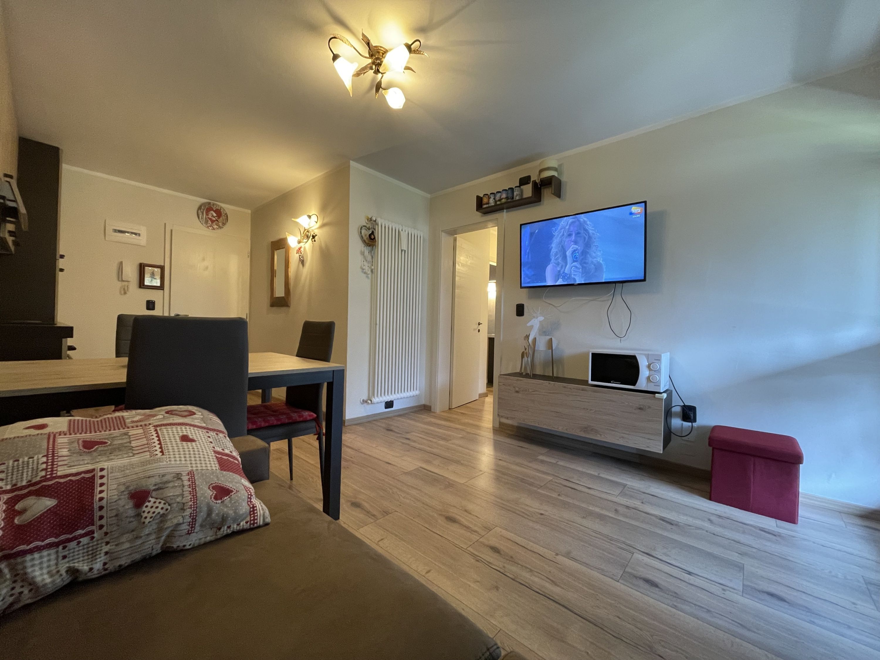 A modern living room with a wooden floor, a dining area with a table and chairs, a television mounted on the wall, and a small red stool.