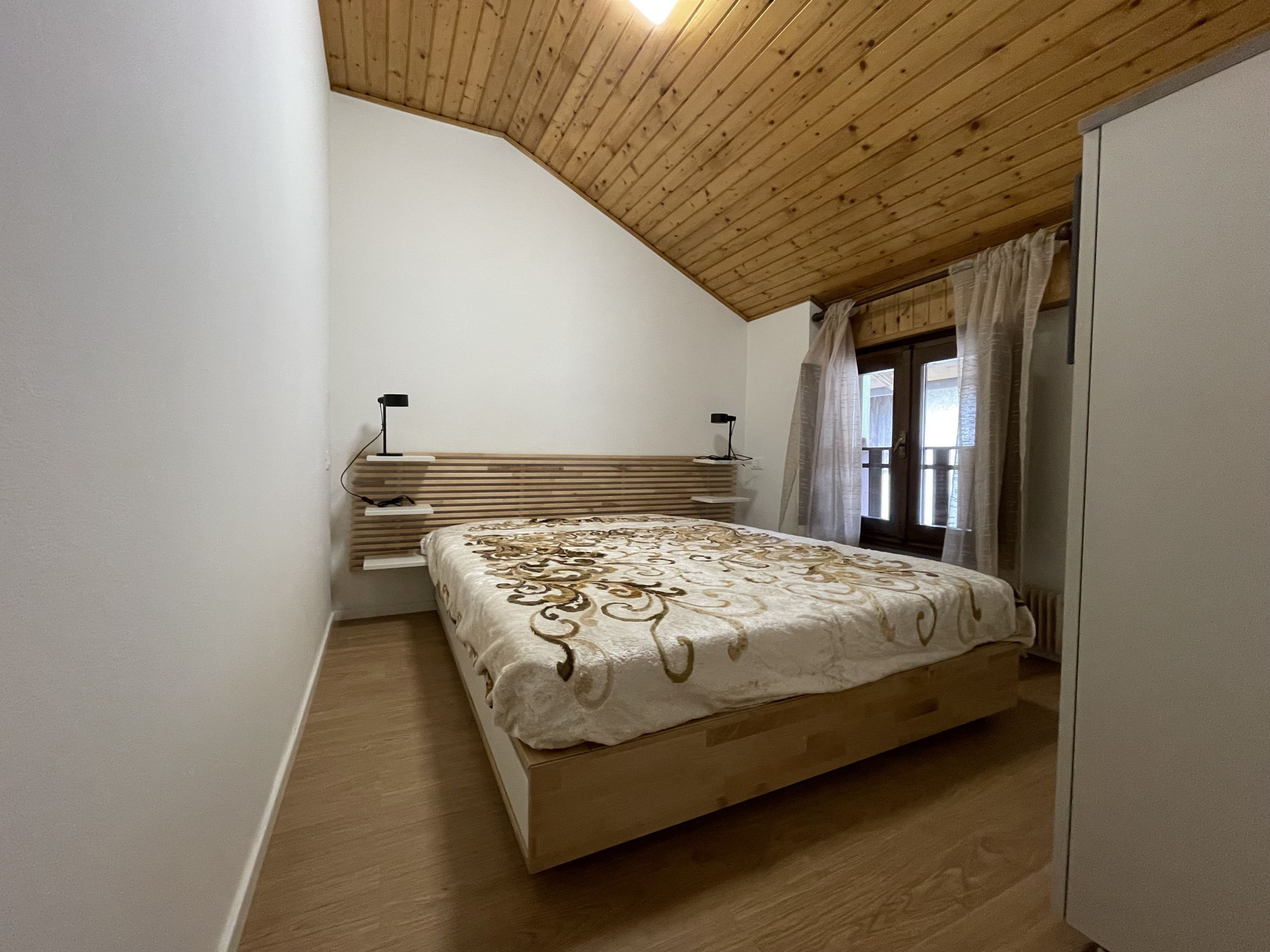 Ein Schlafzimmer mit schrägen Wänden, einem Bett mit gemustertem Überwurf, einem Nachttisch mit Lampe und einem Fenster mit Vorhängen.