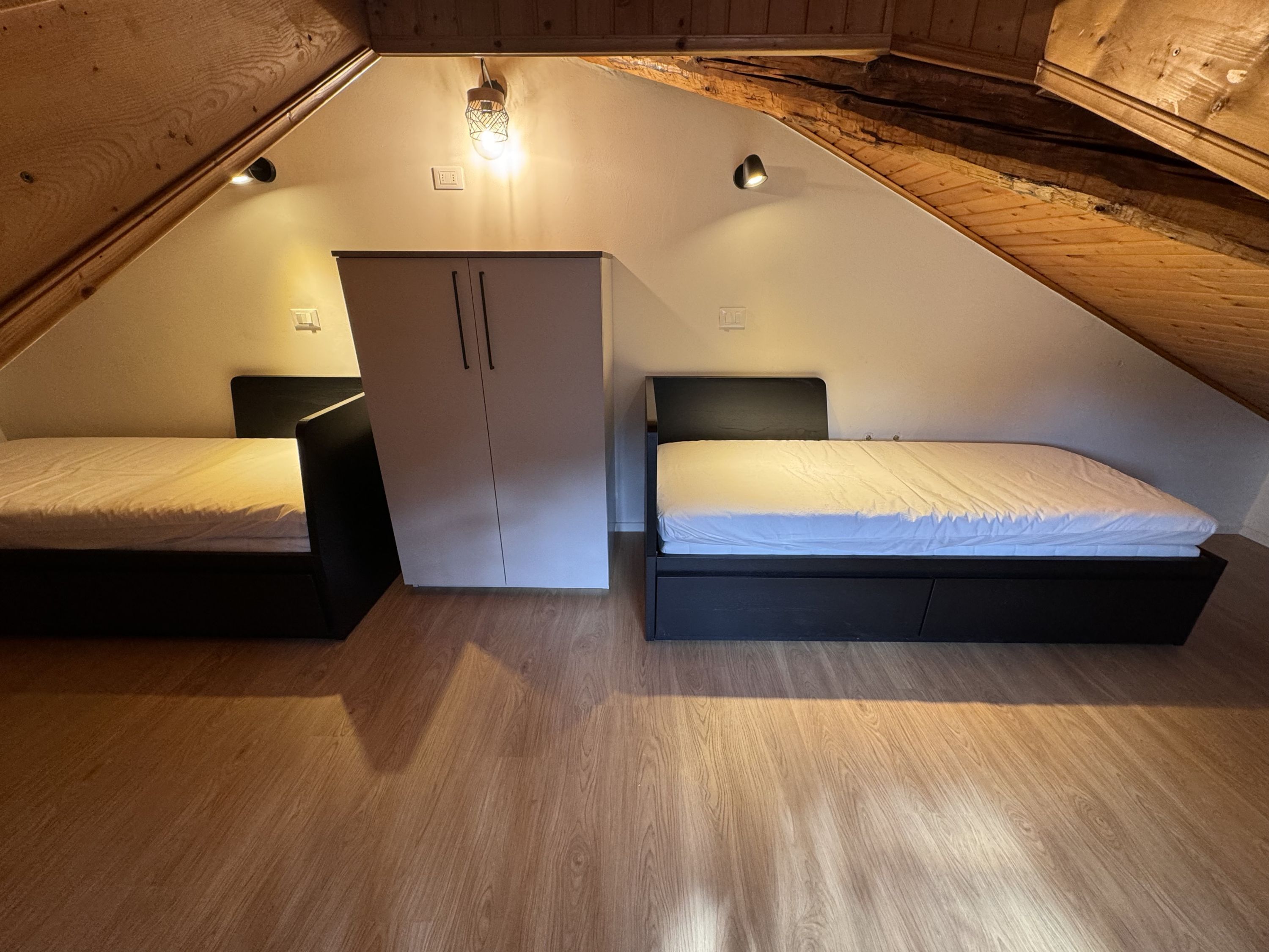 A small, triangular attic bedroom with two beds, a wardrobe, and a sloped ceiling.