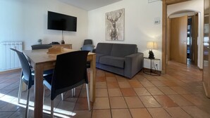 A modern living room with a flat-screen TV, a grey sofa, a dining table with chairs, and a tiled floor.