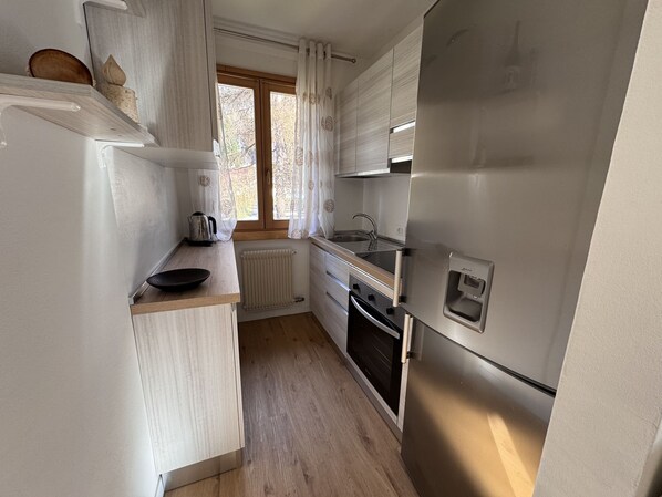 A compact kitchen with white cabinets, a stainless steel refrigerator, a built-in oven, and a window with curtains.