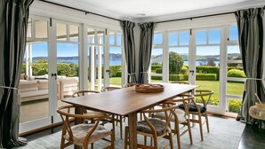 A dining area with a wooden table and chairs, large windows with curtains, and a view of a landscaped garden.